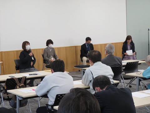 長机に座っている参加者達の前に4名の方が椅子に座り、左端に座っている女性の方がマイクを持って話をしている写真